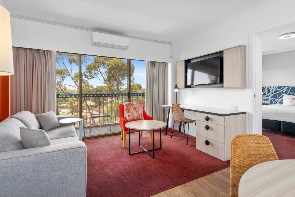 A hotel room at the Plaza Hotel Kalgoorlie, with a red carpet, balcony, sofa, flat-screen TV and work desk